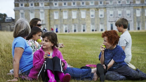 A family group, some eating apples, sitting in the grounds on the grass outside the west front of Petworth House, West Sussex.
